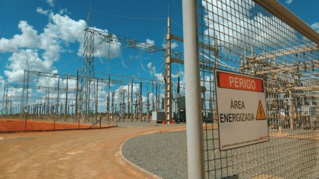 Empty high voltage station with wires and pillars located in sunny sandy area behind metal fence with warning sign