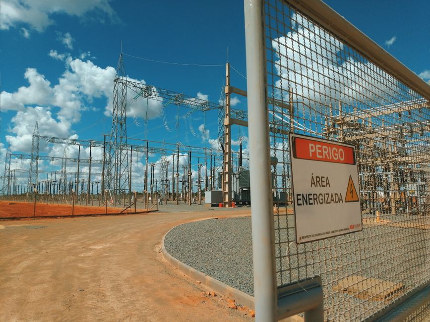 Empty high voltage station with wires and pillars located in sunny sandy area behind metal fence with warning sign