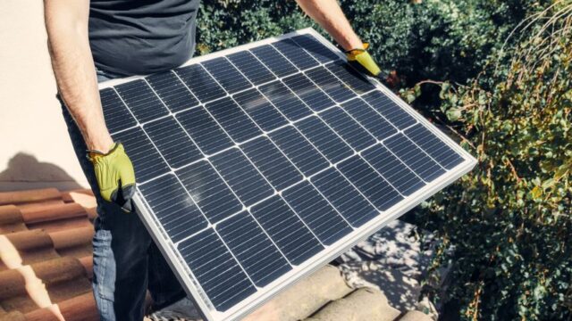 A worker in gloves installs a solar panel on a tiled rooftop, promoting renewable energy.