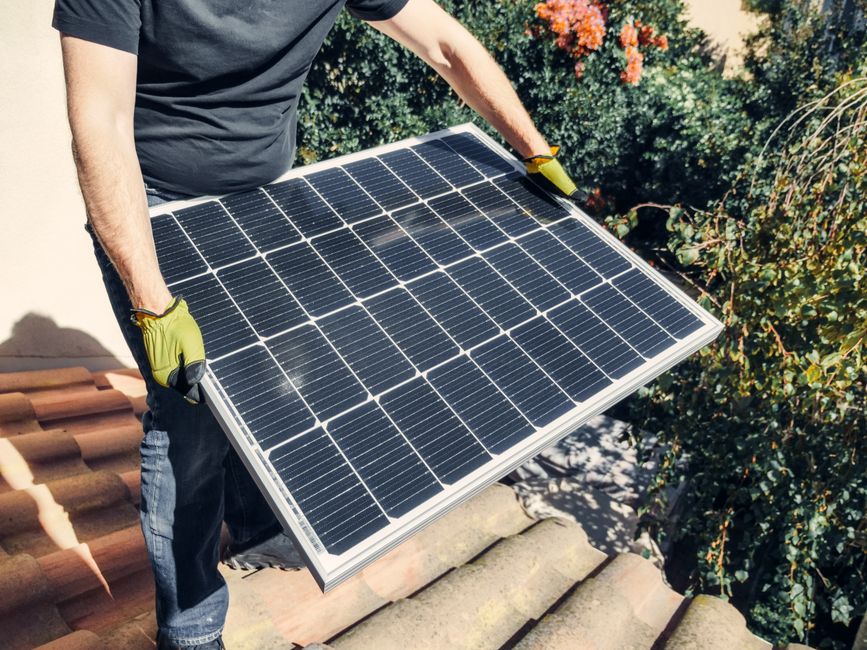 A worker in gloves installs a solar panel on a tiled rooftop, promoting renewable energy.