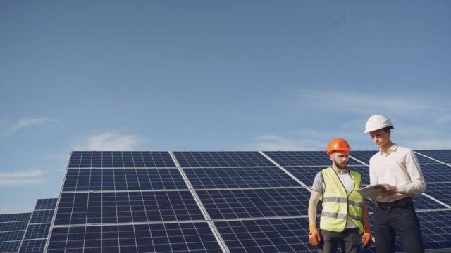 From below of serious young male colleagues in helmets working at solar energy station against cloudless blue sky