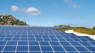 Expansive solar farm with rows of panels under clear blue sky and hills in the background.