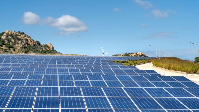 Expansive solar farm with rows of panels under clear blue sky and hills in the background.