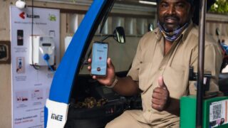 South Asian man using electric vehicle charging station in India, holding smartphone with app.