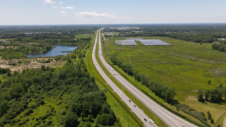 Aerial view of a highway alongside a solar farm and greenery, captured on a bright summer day.