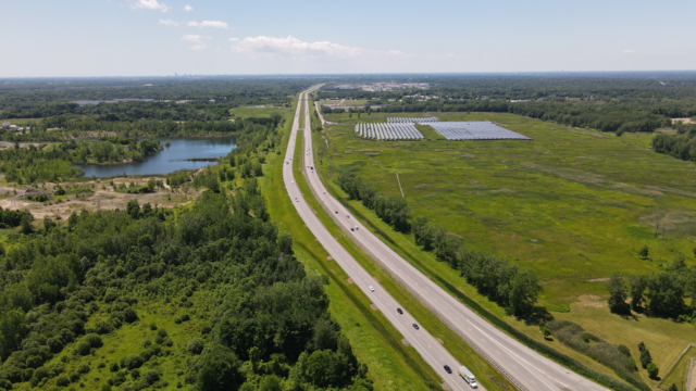 Aerial view of a highway alongside a solar farm and greenery, captured on a bright summer day.