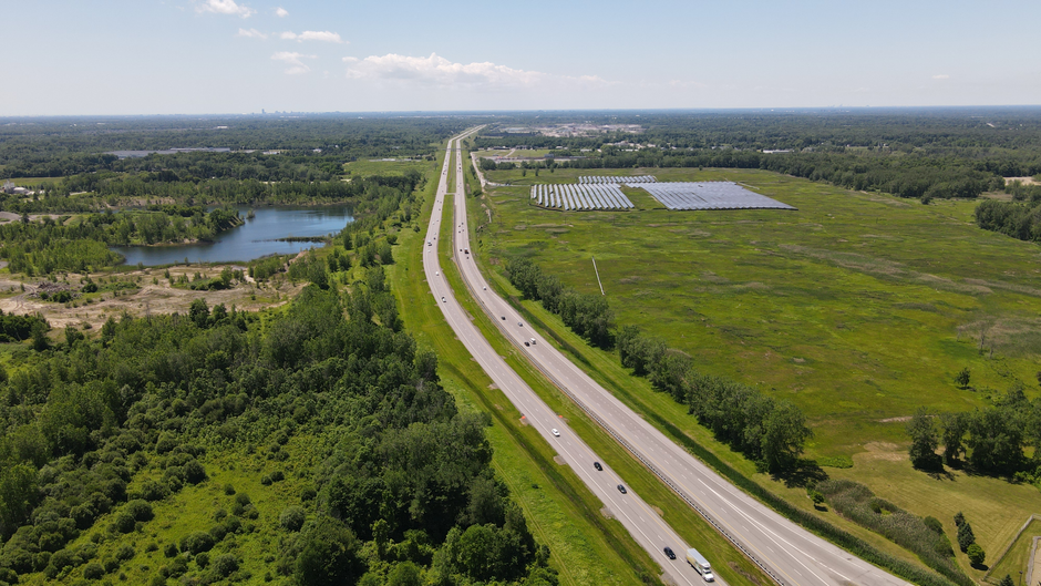 Aerial view of a highway alongside a solar farm and greenery, captured on a bright summer day.