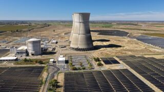 Aerial view of a nuclear cooling tower surrounded by solar panel fields in a rural landscape.