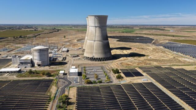 Aerial view of a nuclear cooling tower surrounded by solar panel fields in a rural landscape.