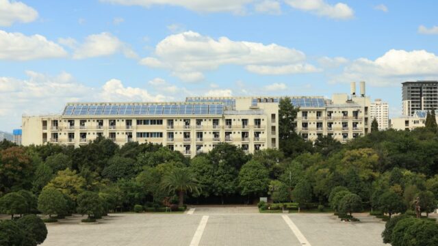 A modern building with solar panels, surrounded by lush greenery, under a blue sky with clouds.