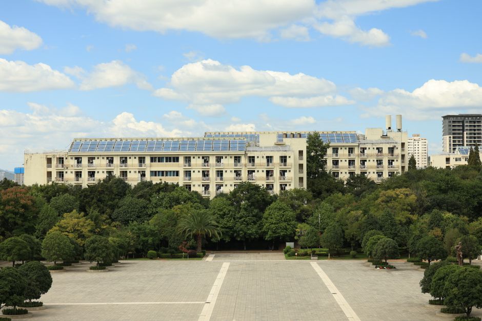 A modern building with solar panels, surrounded by lush greenery, under a blue sky with clouds.
