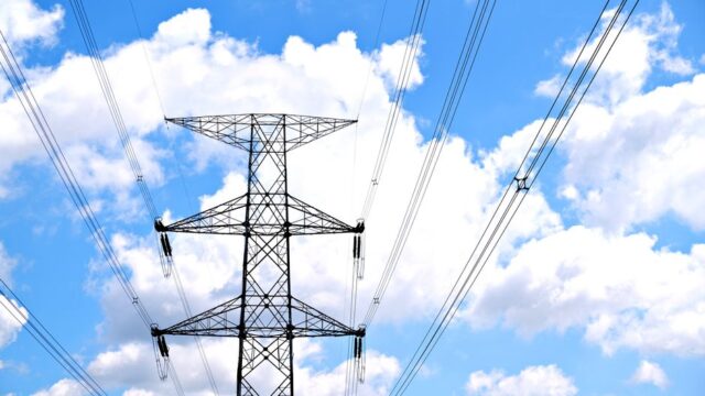 A view of high voltage power lines against a bright blue sky with fluffy clouds.
