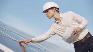 Low angle of young male inspector with documents in formal wear and hardhat standing against solar panels and checking details of construction