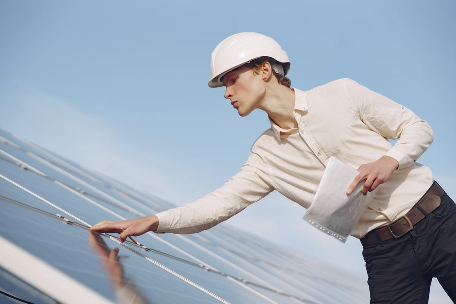 Low angle of young male inspector with documents in formal wear and hardhat standing against solar panels and checking details of construction