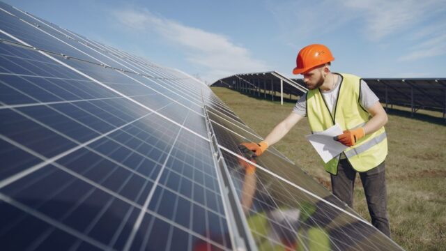 Young bearded male engineer in protective helmet and heat resistant gloves touching solar panel while standing on field with papers on sunny day